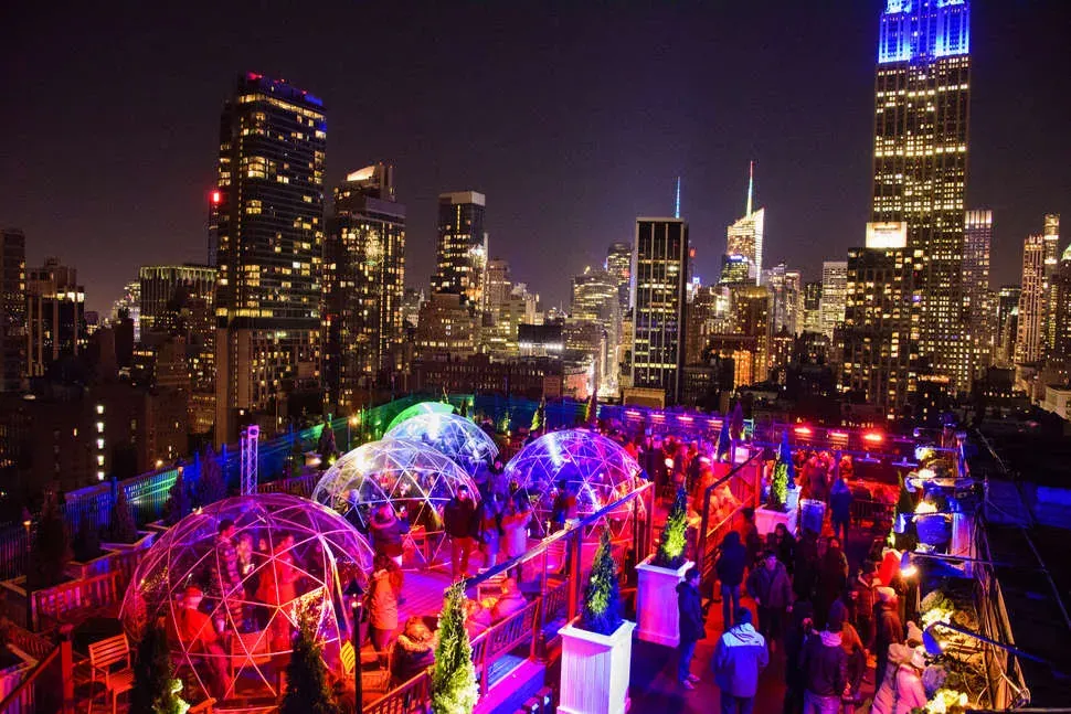 Clear dome igloos and crowds on the 230 Fifth rooftop at night with the Empire State Building lit behind