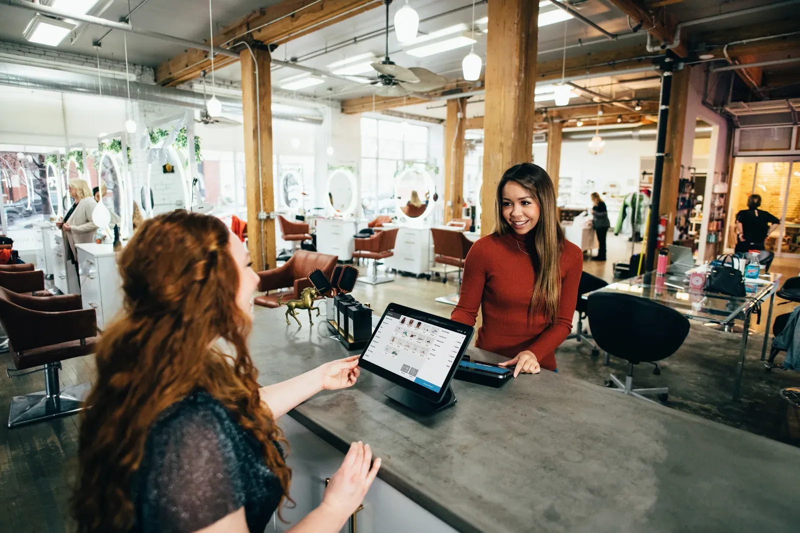Client paying at a salon front desk on a tablet point-of-sale system as a smiling receptionist helps her