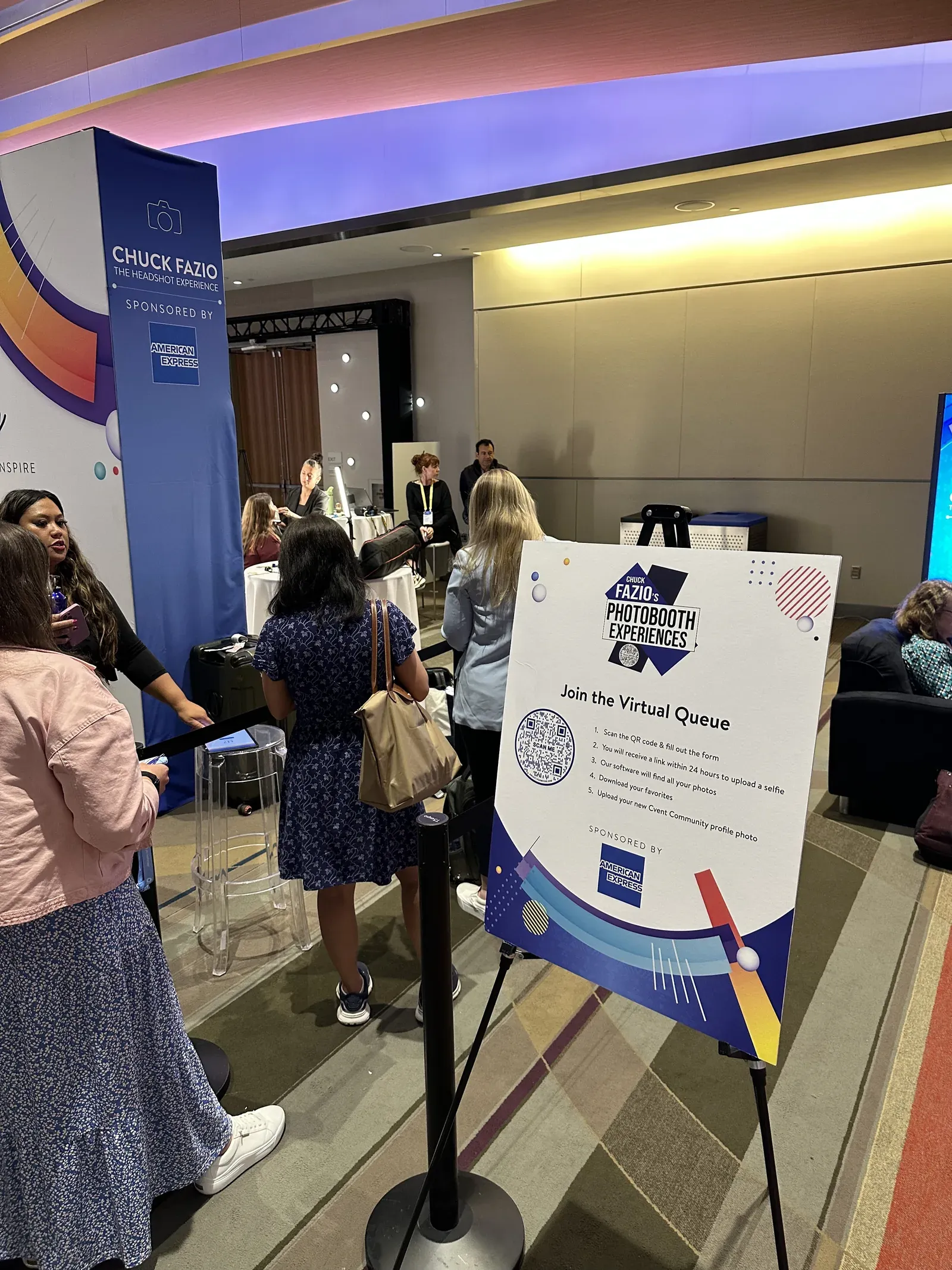 Attendees lining up at a sponsored headshot booth beside an easel sign with a QR code inviting them to join the virtual queue