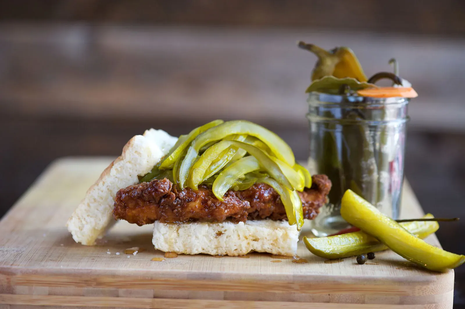Jacob’s Pickles fried chicken biscuit sandwich piled with pickles next to a jar of pickled peppers