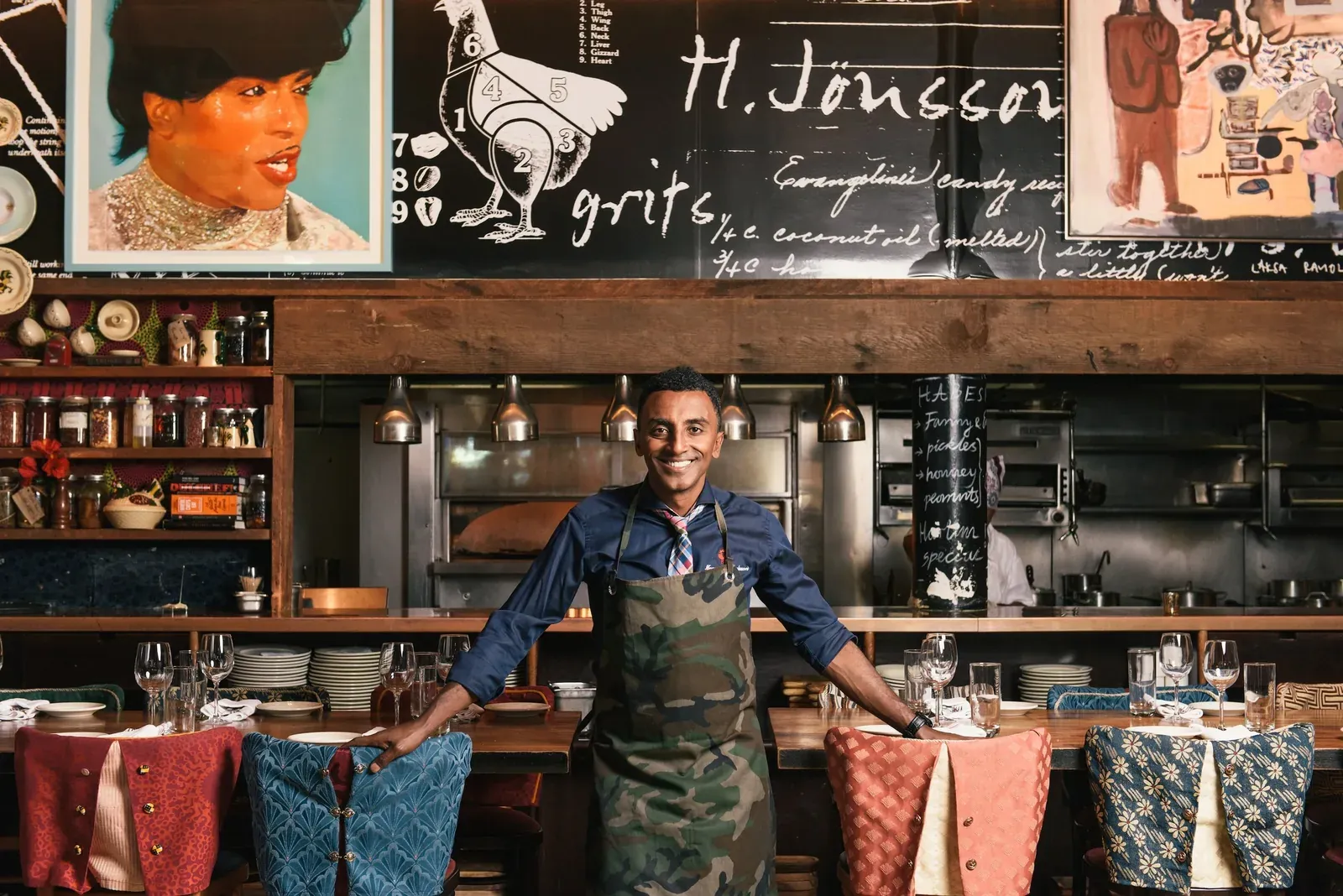 Chef Marcus Samuelsson smiling in a camo apron inside the Red Rooster dining room in front of the open kitchen