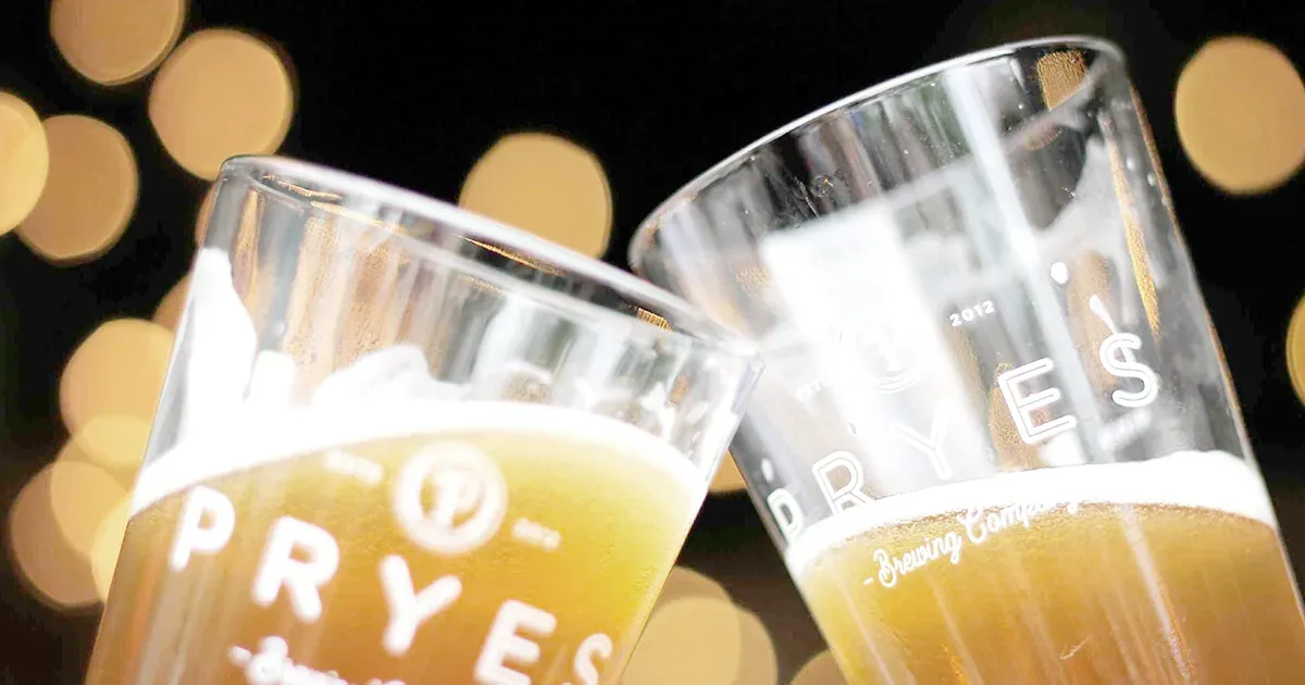 Two Pryes Brewing Company pint glasses of beer clinking against a background of warm out-of-focus lights