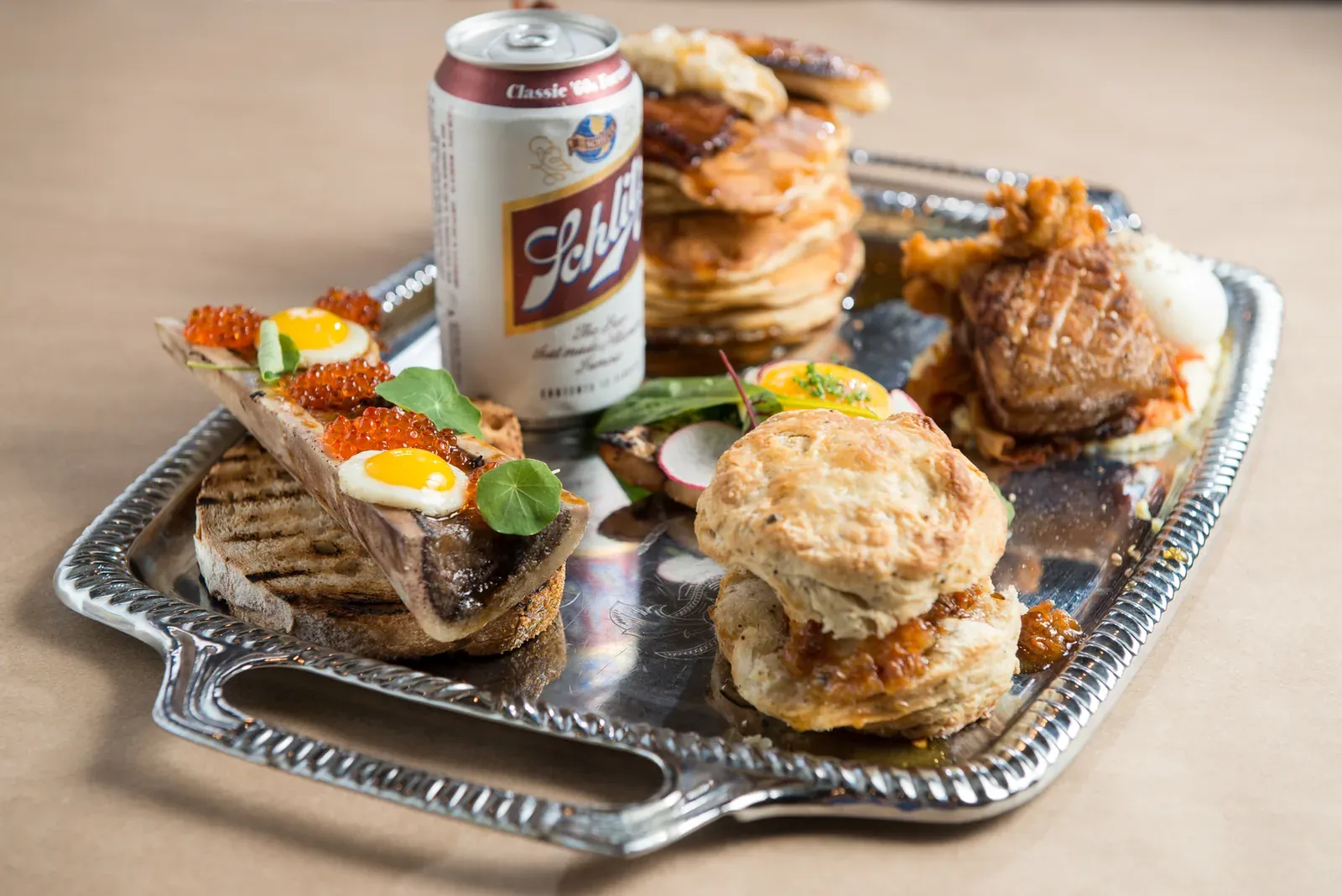 One Eared Stag brunch platter with bone marrow toast, a biscuit, a pancake stack, and a Schlitz can on a silver tray