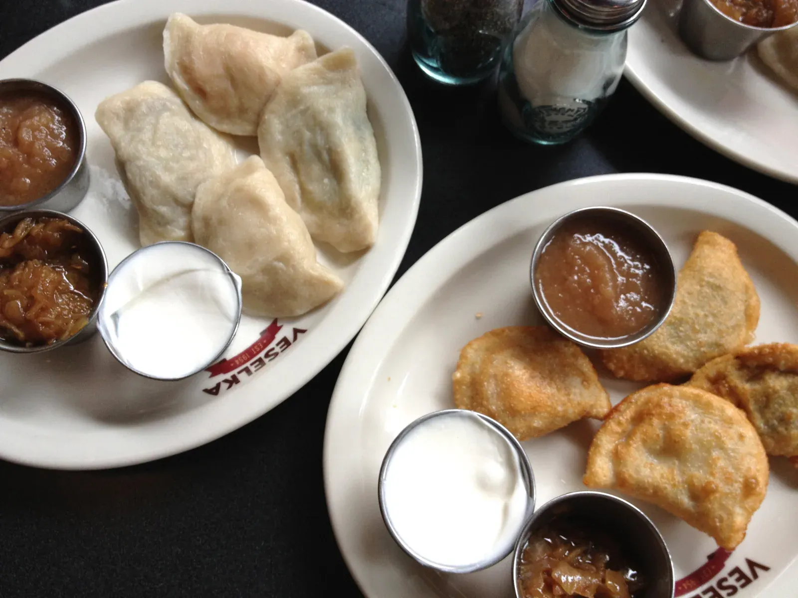 Two Veselka plates of boiled and fried pierogi with sides of sour cream, applesauce, and fried onions