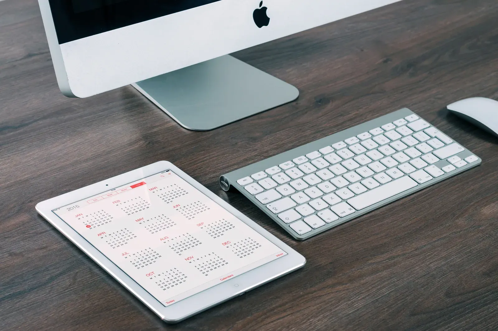 Tablet showing a yearly calendar on a desk next to a keyboard and an iMac