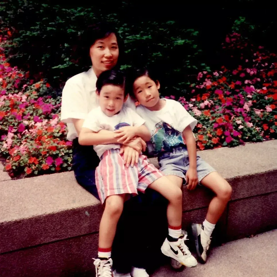 A childhood photo of James and John Yi sitting with their mother, Anna, in front of a flower bed