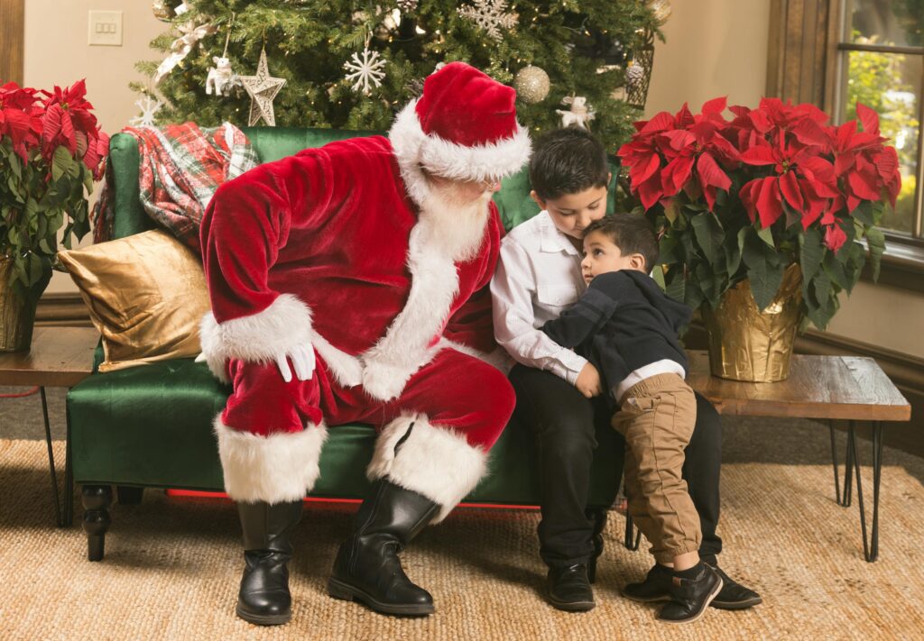 Santa Claus shares a joyful moment with two children by a Christmas tree indoors.